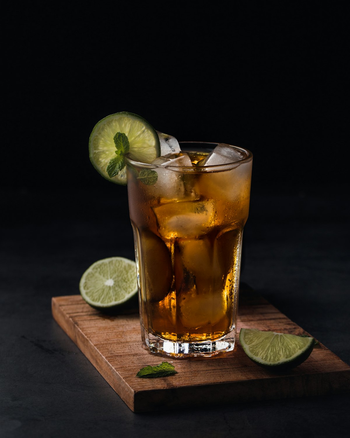 Tall glass of chilled amber drink with ice, lime, and mint on a wooden board against a dark background